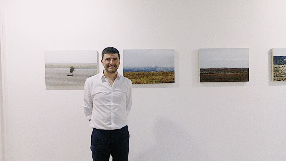 Un hombre con camisa blanca de pie frente a fotografías de paisajes en una pared de galería. Las fotografías representan varias escenas naturales, incluyendo campos y horizontes distantes.