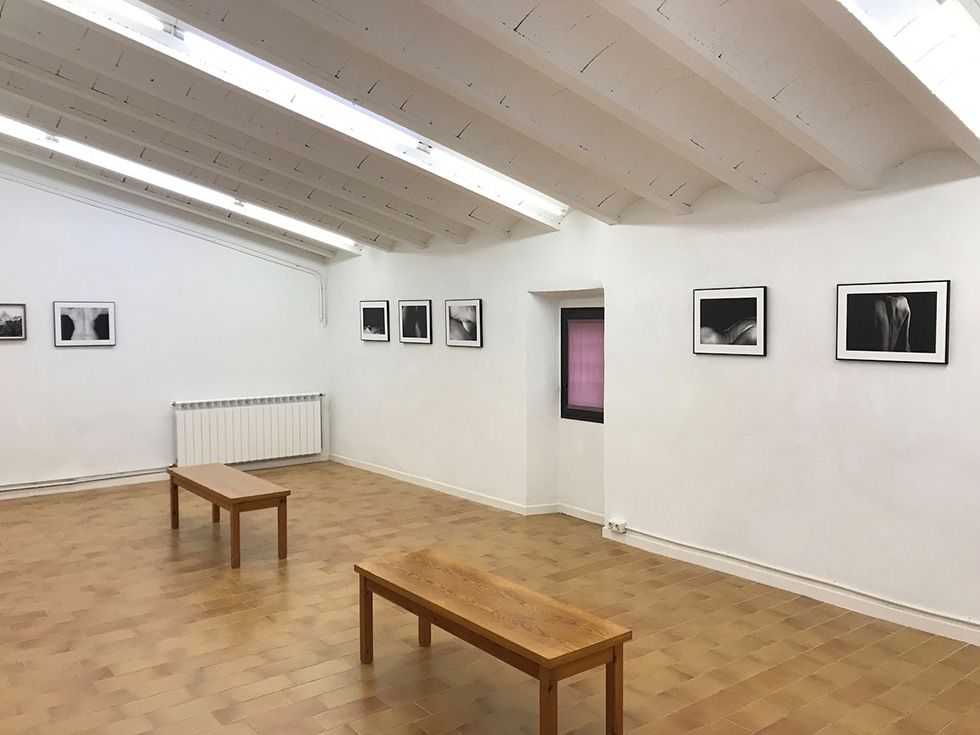 Una sala de galeria minimalista amb fotografies en blanc i negre en parets blanques, dos bancs de fusta al centre i una petita finestra en una paret.