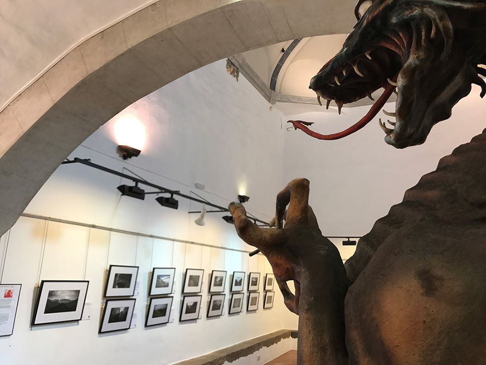 Exposición en galería con fotografías enmarcadas y en primer plano una dramática efigie de un dragón legendario catalán.