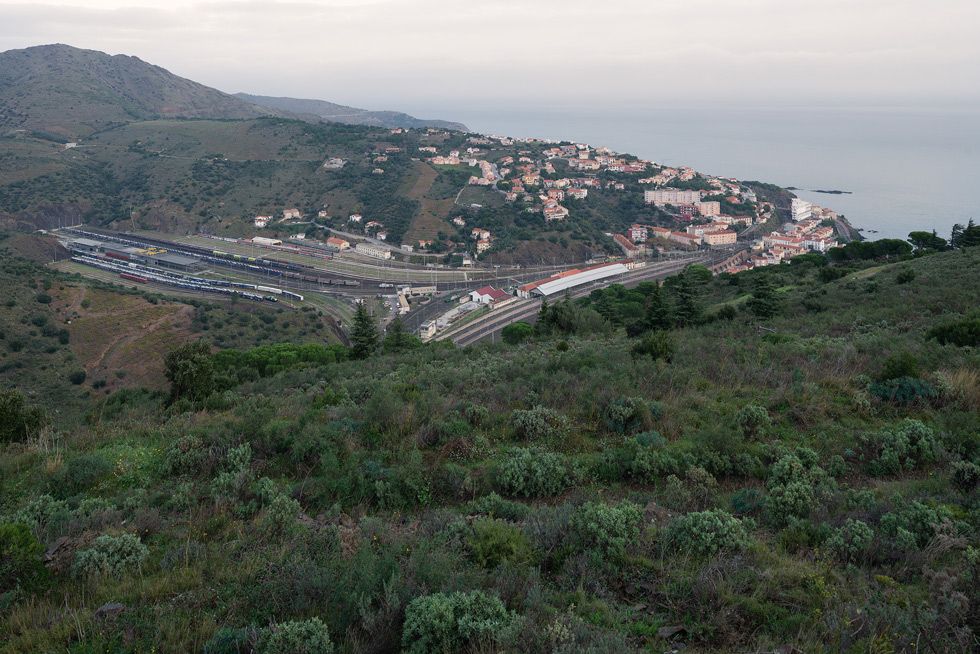 Vista d'un poble costaner amb una gran estació de tren amb múltiples vies sense sortida que travessen una vall i cases escampades pel turó, estenent-se cap al mar.