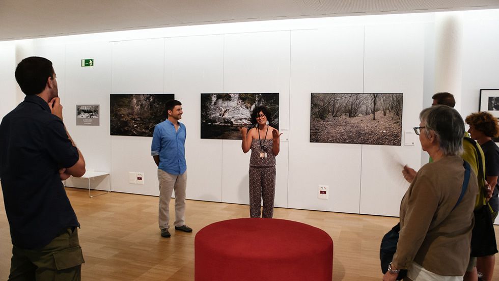 Un grup de persones escoltant un orador en una galeria d'art, amb grans fotografies de paisatges exposades a la paret darrere d'ells.