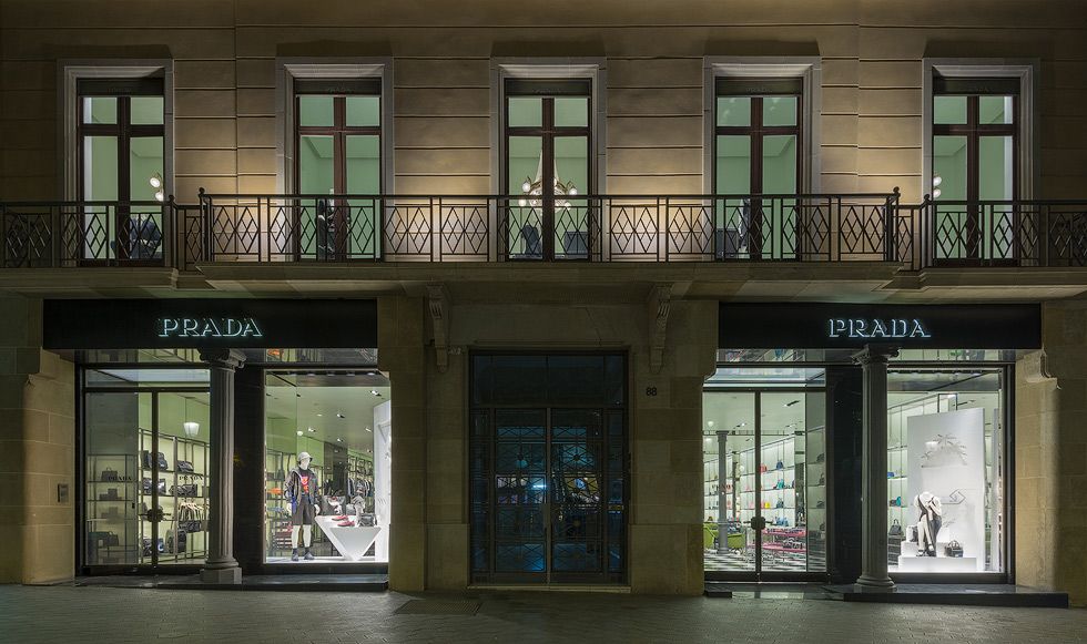 Exterior d'una botiga Prada de nit, mostrant aparadors brillantment il·luminats amb articles de luxe i maniquins, emmarcats per una façana d'edifici clàssic.