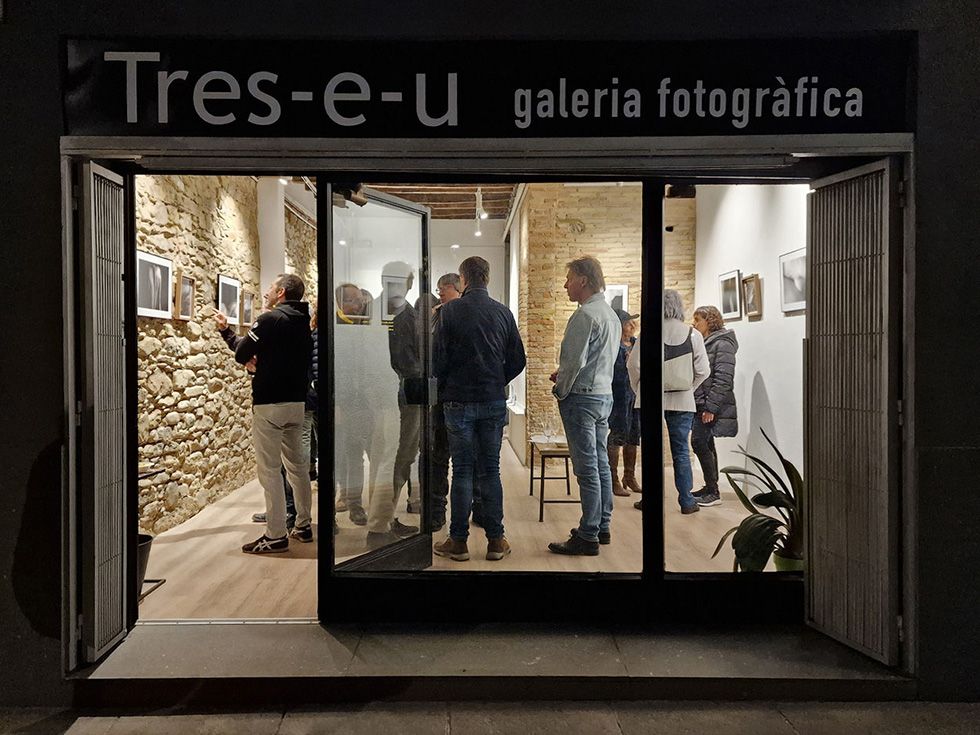 Persones observant fotografies a la galeria de fotografia Tres-e-u, vistes des del carrer a través de l'entrada principal de la galeria.
