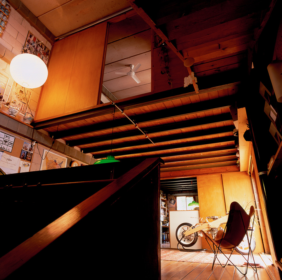 Low-angle interior view of a warm, eclectic space with exposed steel beams, sliding wooden panels opening into a loft area with a ceiling fan. In the ground floor, a wood motorcycle prototype is visible in the background.