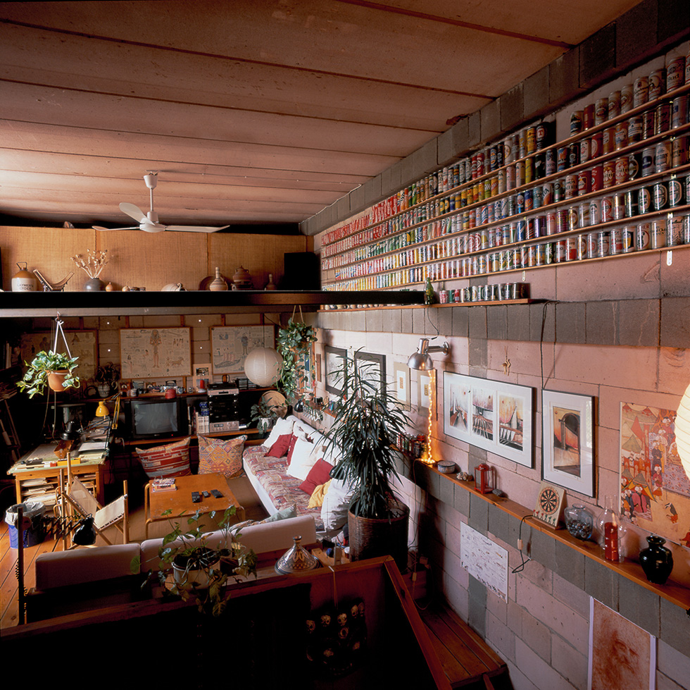 Cozy living space with an heterogeneous collection of items, including a large display of cans on the wall, various artworks, plants, and a mix of modern and vintage furniture.