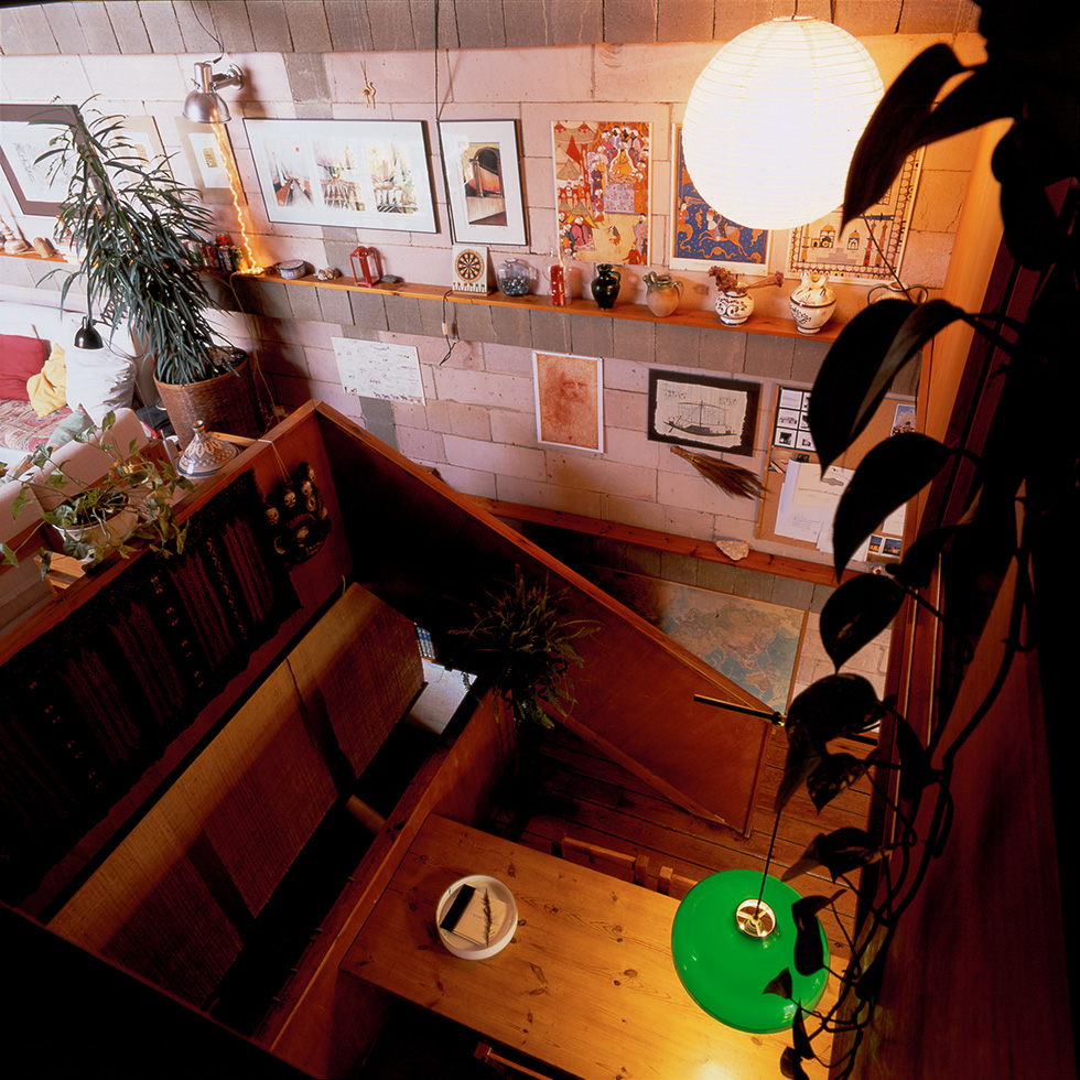 High-angle view of a cozy living area with a wooden staircase, a green pendant light, and a wall adorned with framed art and decorative items. Below you can see a large wooden table.