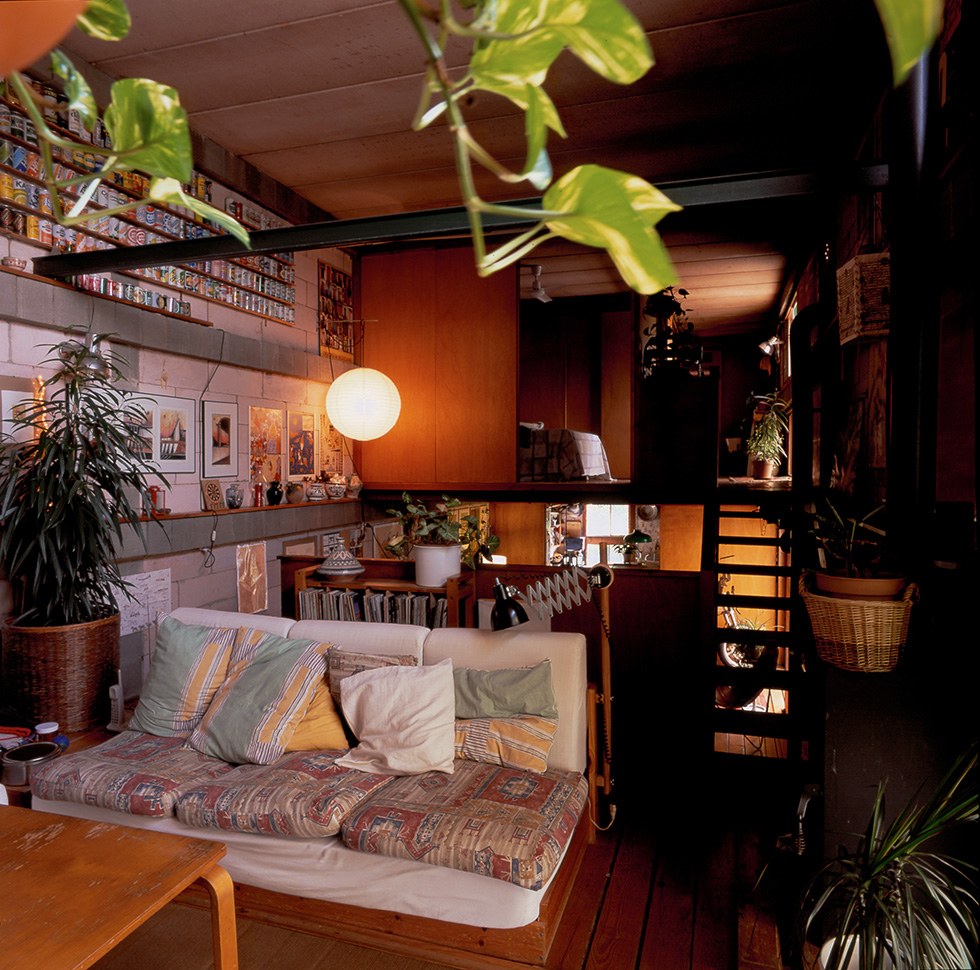 Cozy living room with a colorful sofa, wooden furniture, indoor plants, and a white pendant light, featuring a staircase and a wall decorated with art and collectibles.