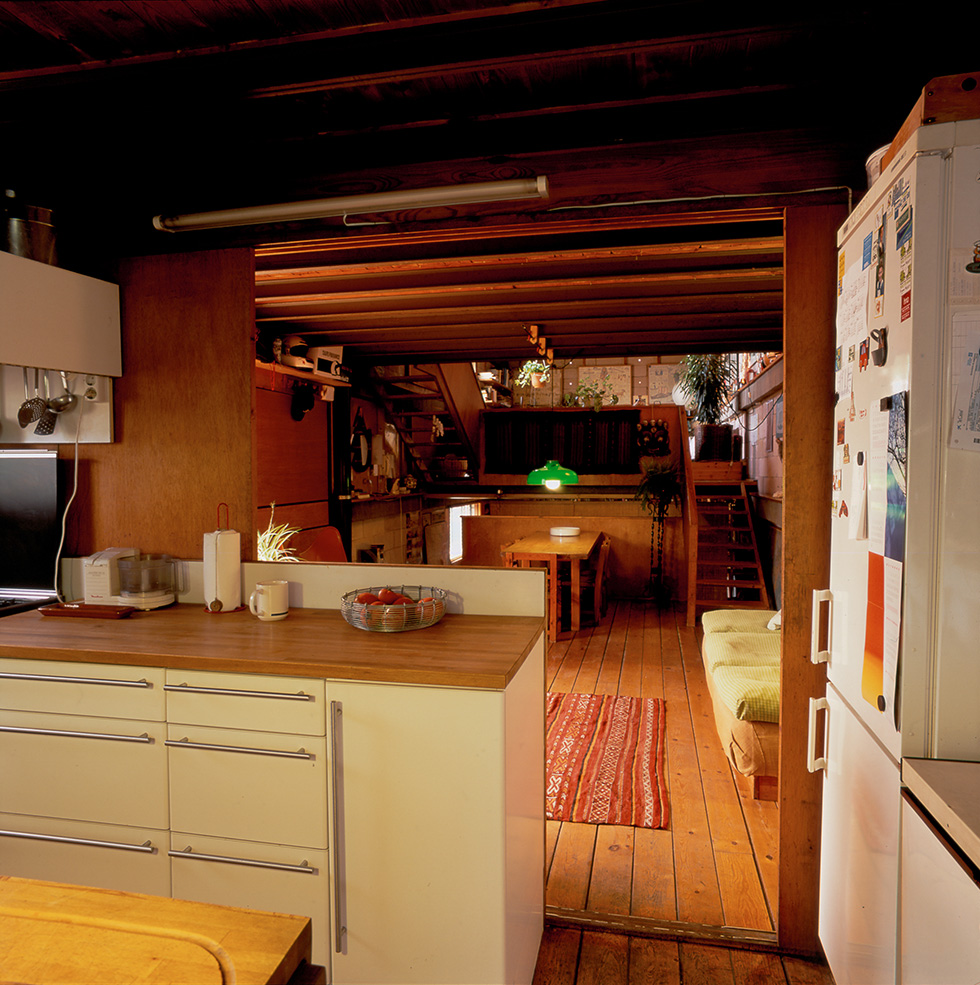 Cozy kitchen with a wooden floor and ceiling, white cabinets, leading to a dining area with a colorful rug, a wooden table, green pendant light, and an open air wooden staircase in the background.