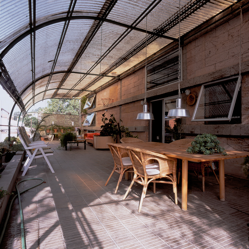 Covered outdoor patio with a translucent coorrugated polycarbonate arched roof, wooden dining table with wicker chairs, plants, and natural light streaming through.