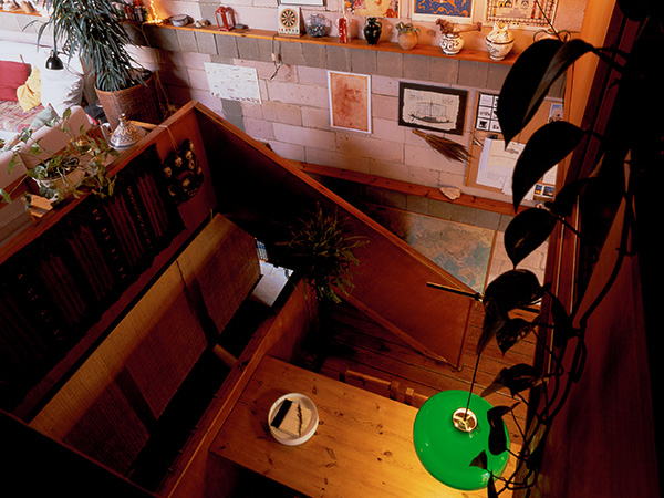 High-angle view of a cozy living area with a wooden staircase, a green pendant light, and a wall adorned with framed art and decorative items. Below you can see a large wooden table.