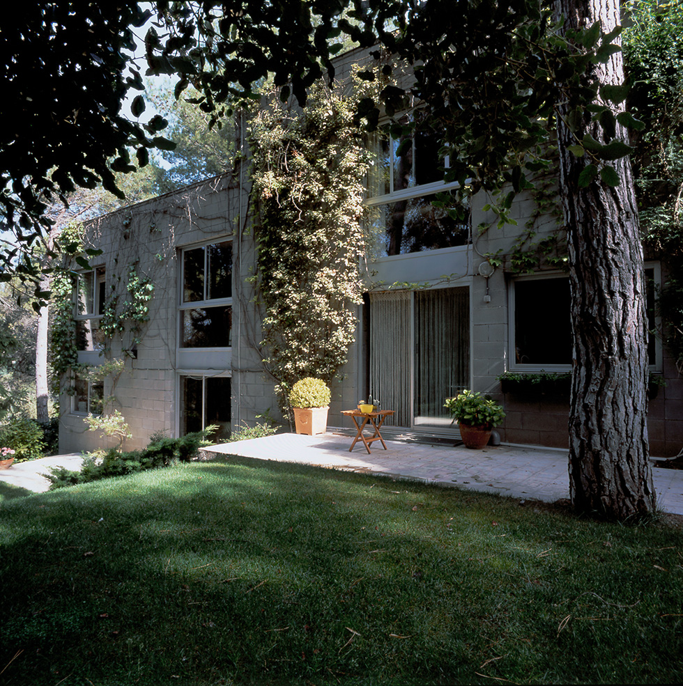 Two-story terraced house exterior with large windows, ivy-covered walls, a paved area with potted plants and a small table, all surrounded by large trees and a lawn.