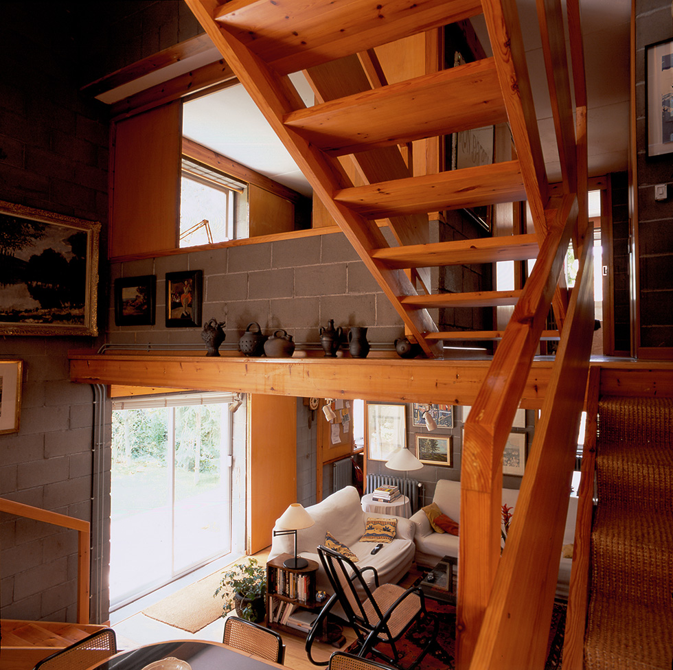 Interior view showing a wooden staircase, a cozy living room with white sofas, a rocking chair, a rug, and decorative items on shelves and walls. The room has concrete block walls and a large sliding door letting in natural light.