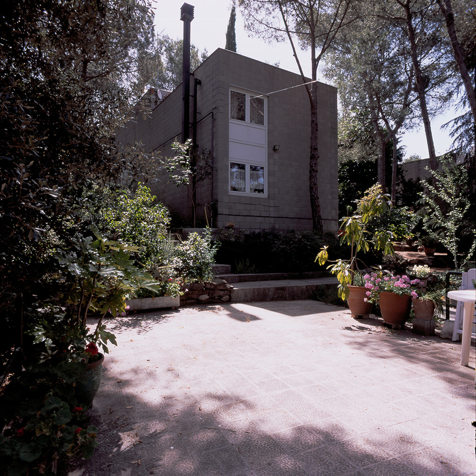 Exterior view of a concrete block house with a garden patio, surrounded by potted plants and trees, creating a serene and shaded outdoor space.