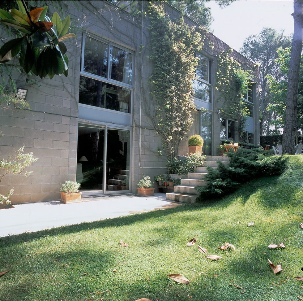 View of a terraced concrete block house with large windows, surrounded by lush greenery, a grassy lawn, and a staircase leading to an elevated paved area adorned with potted plants.