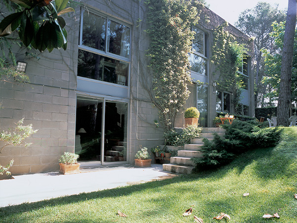 View of a terraced concrete block house with large windows, surrounded by lush greenery, a grassy lawn, and a staircase leading to an elevated paved area adorned with potted plants.