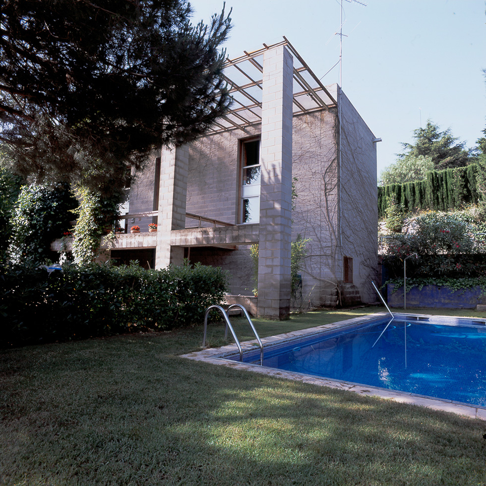 Exterior view of a modern concrete block house with a large pool in the garden, surrounded by greenery and trees.