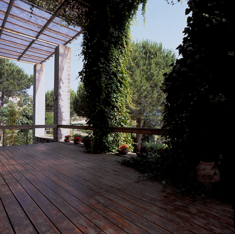 Wooden deck with ivy-covered concrete block pergola, overlooking a garden with pine trees and potted flowers on the floor.