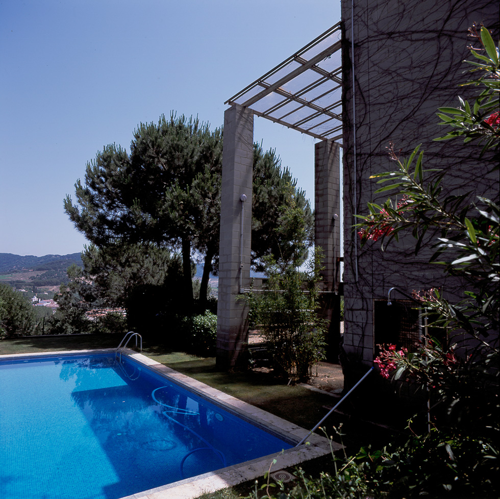 Outdoor swimming pool beside a concrete block house, surrounded by trees, greenery and oleander flowers, with a view of distant hills.