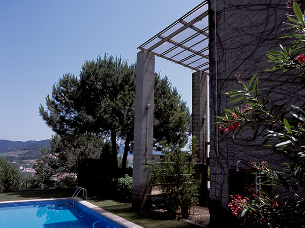 Outdoor swimming pool beside a concrete block house, surrounded by trees, greenery and oleander flowers, with a view of distant hills.