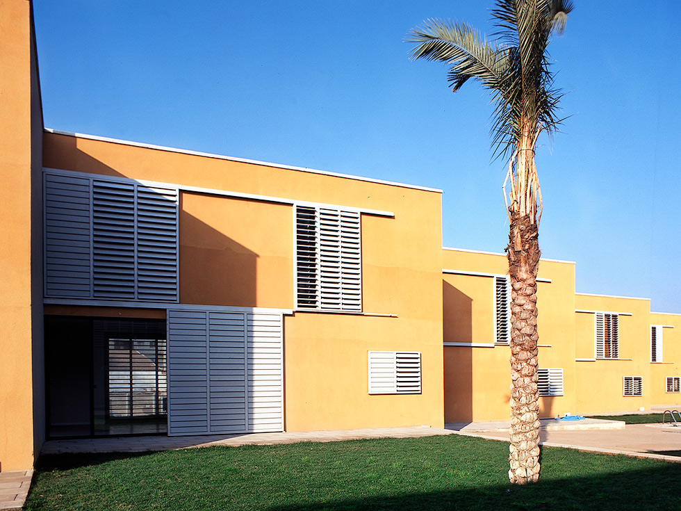 A cluster of modern, flat-roofed buildings with yellow facades and large louvered windows, set against a bright blue sky and featuring a palm tree in the foreground.