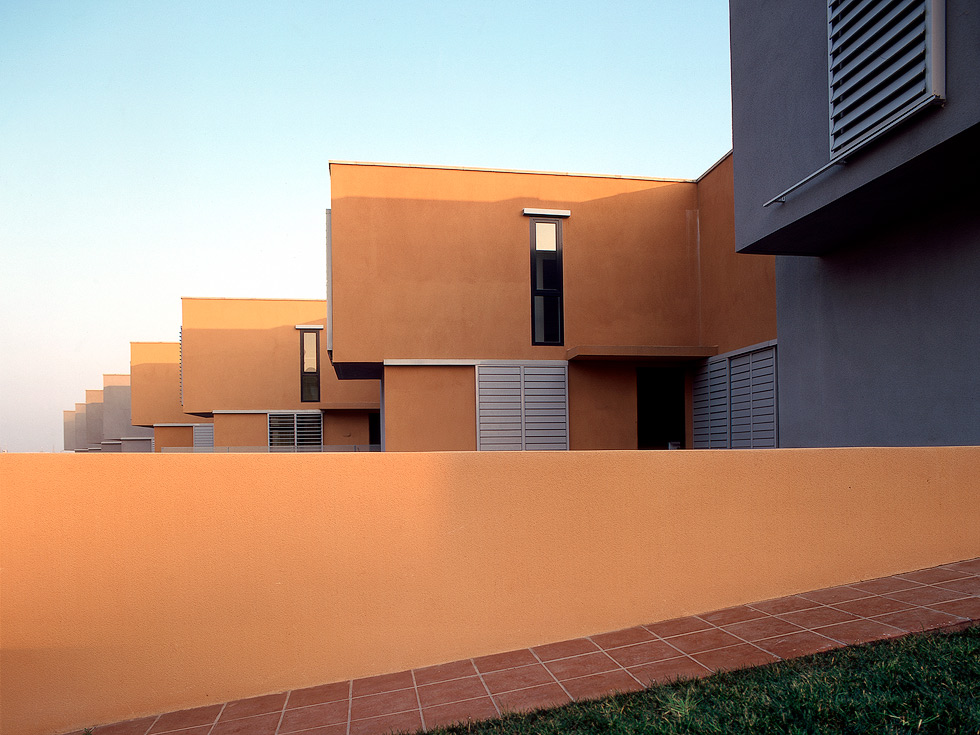 Modern row houses with flat roofs and large louvered windows, painted in warm tones, with a terracotta-tiled walkway and a grassy area in the foreground.