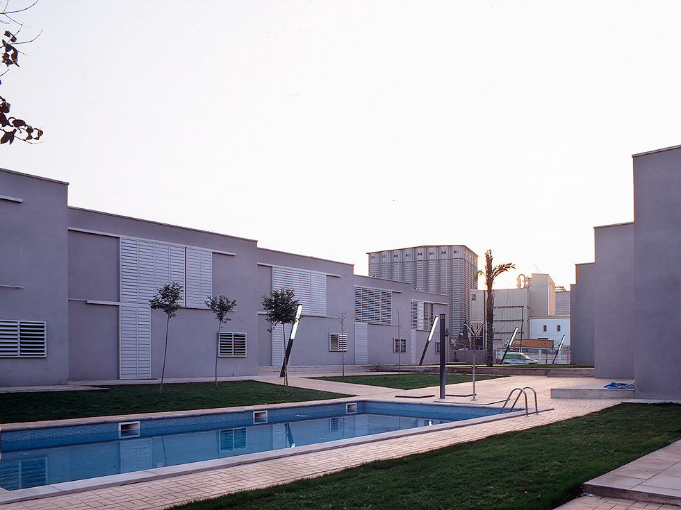 Modern residential complex with flat-roofed buildings, louvered windows, a central swimming pool, and landscaped grassy areas.