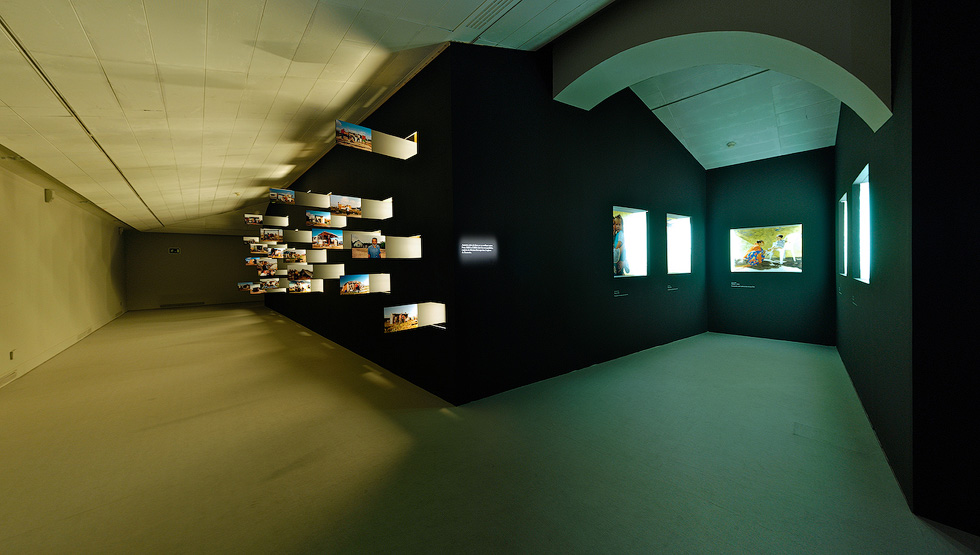 A dimly lit exhibition room featuring multiple illuminated display boxes and shelves showcasing various photos and objects on the walls.