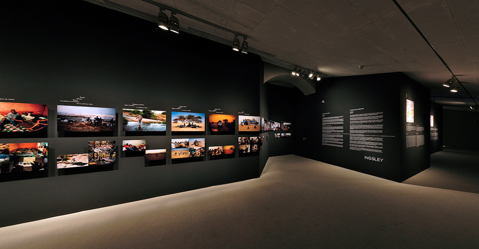 A gallery showcasing a series of photographs and descriptive text panels on dark walls, illuminated by focused spotlights.