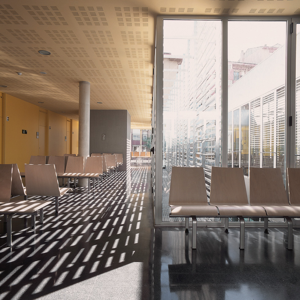 A modern waiting area with light wooden chairs, large windows, yellow walls and ceiling, creating a bright and airy atmosphere.