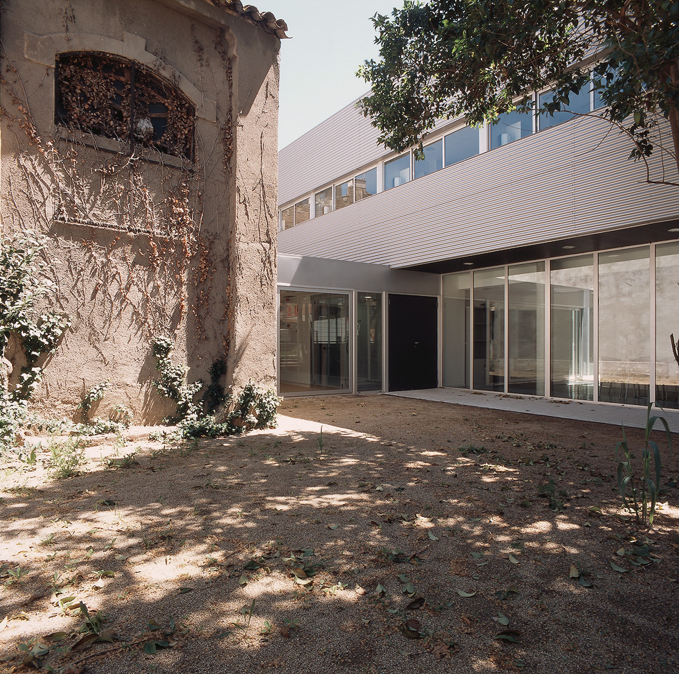 A courtyard blending old and new architecture, with a historic building on the left and a modern glass structure on the right, surrounded by greenery and shaded trees.