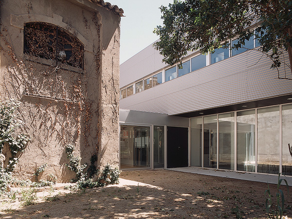 A courtyard blending old and new architecture, with a historic building on the left and a modern glass structure on the right, surrounded by greenery and shaded trees.