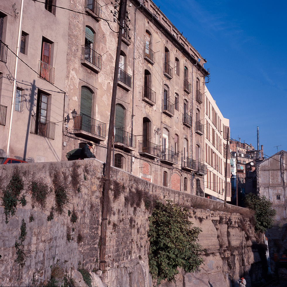 Old residential building with a weathered facade, featuring balconies and set on a stone embankment. In the background a new residential building following the organic street pattern