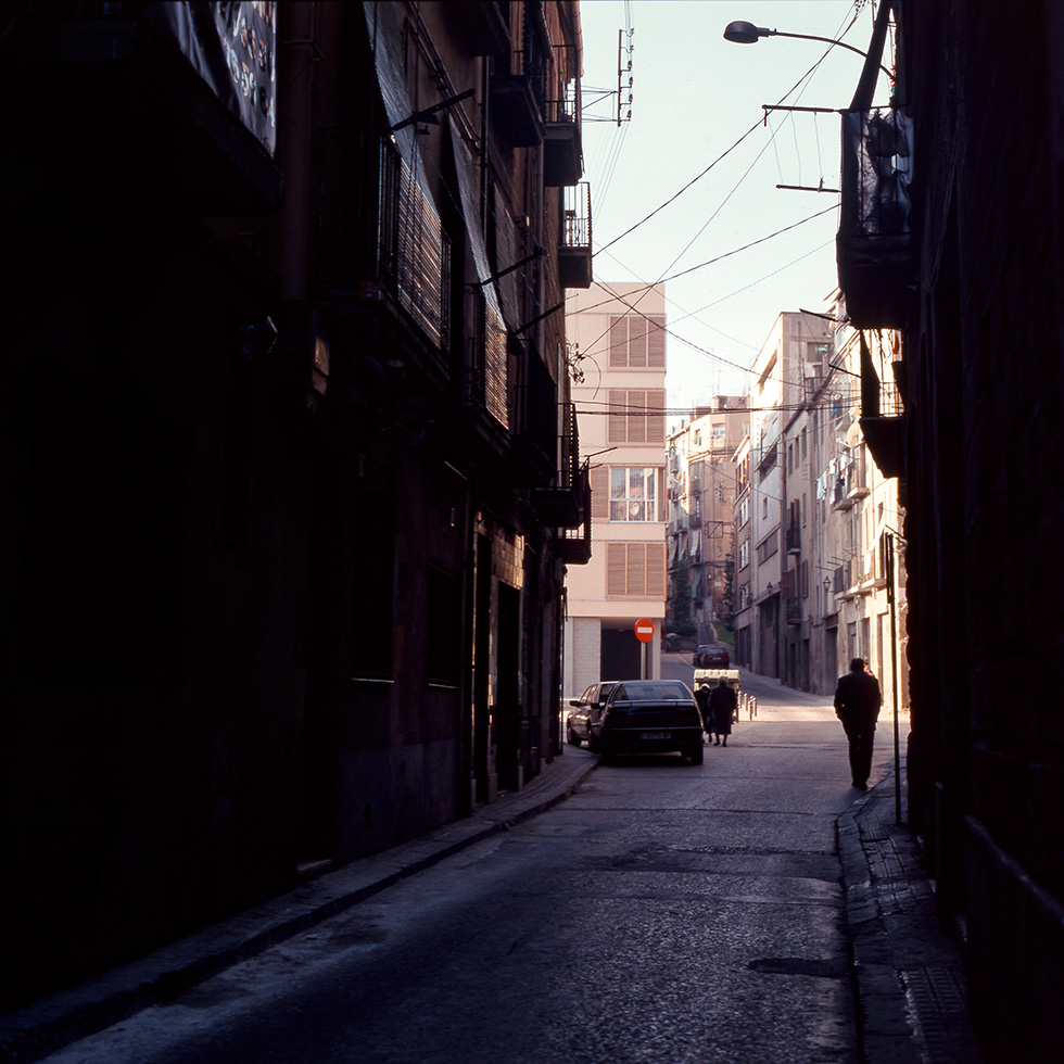 A narrow alleyway with shadowed buildings on both sides, leading to a brighter plaza and a new residential building in the background with a few pedestrians and parked cars.