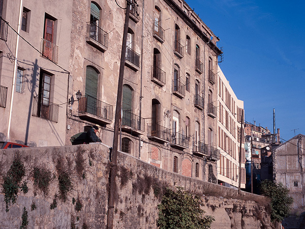 Old residential building with a weathered facade, featuring balconies and set on a stone embankment. In the background a new residential building following the organic street pattern