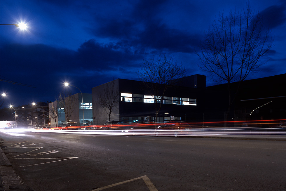 Nighttime street scene with a modern building in the background, car light trails, and streetlights illuminating the area.