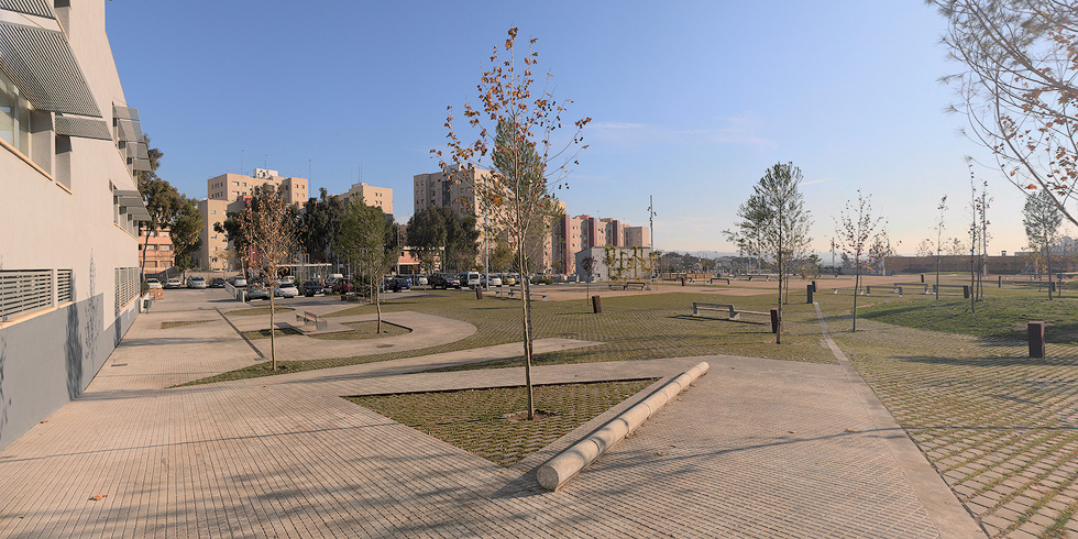 A park with paved walkways, young trees, grassy areas, and benches, set against a backdrop of residential buildings.