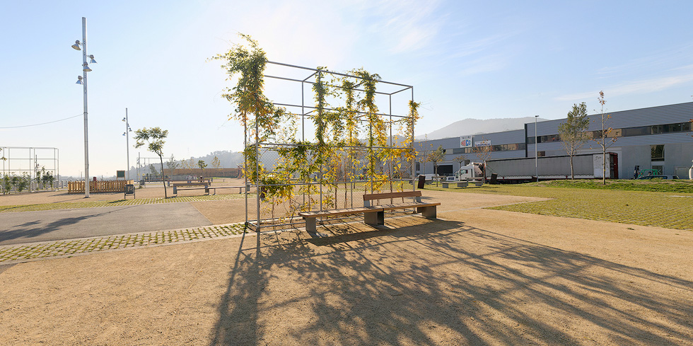 A park with metal trellises supporting climbing plants, benches, pathways, and young trees, with a building and mountains in the background.