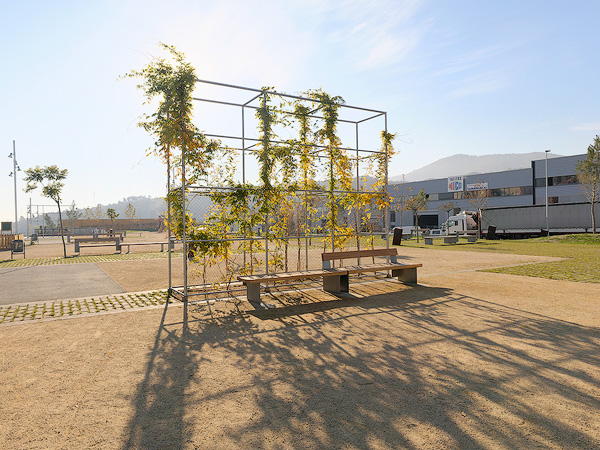 A park with metal trellises supporting climbing plants, benches, pathways, and young trees, with a building and mountains in the background.