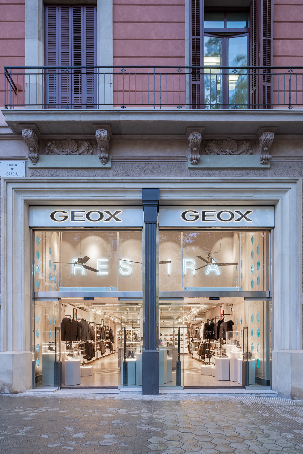 Front view of a Geox store on Passeig de Gracia in Barcelona, with large glass windows displaying the interior merchandise and a balcony above.