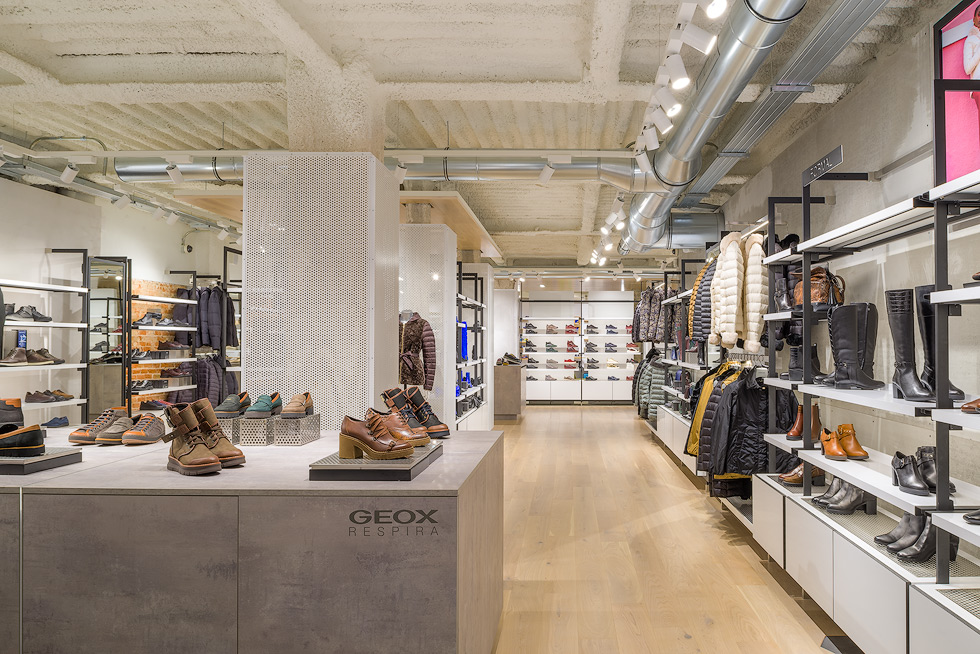 Interior of Geox store displaying a variety of shoes and outerwear, with a modern industrial ceiling design.