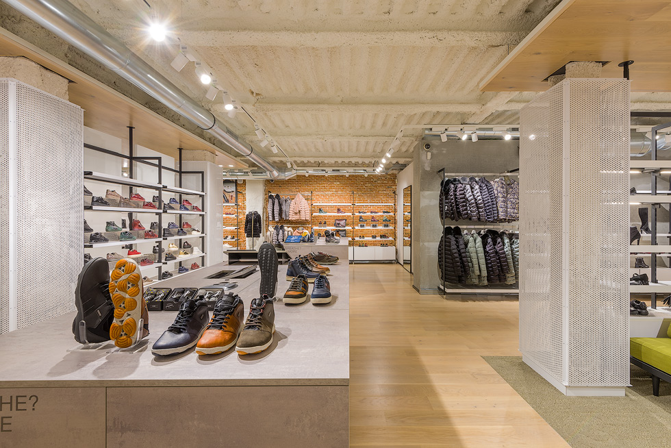 Interior of Geox store showcasing an assortment of shoes on display tables, shelves with shoes, and racks of jackets, with a modern industrial ceiling and exposed brick wall in the background.