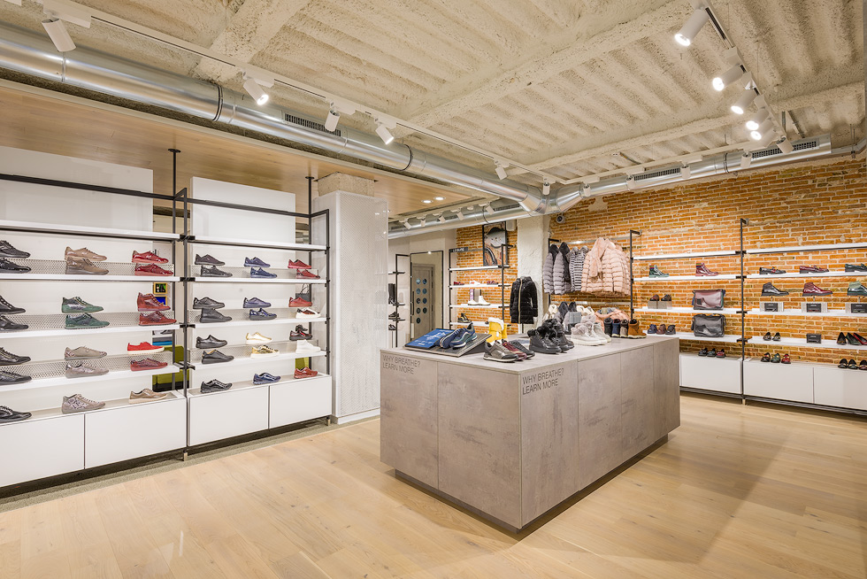 Interior of Geox store, displaying colorful sneakers on shelves and various winter jackets on racks against an exposed brick wall, under modern lighting and exposed ductwork.