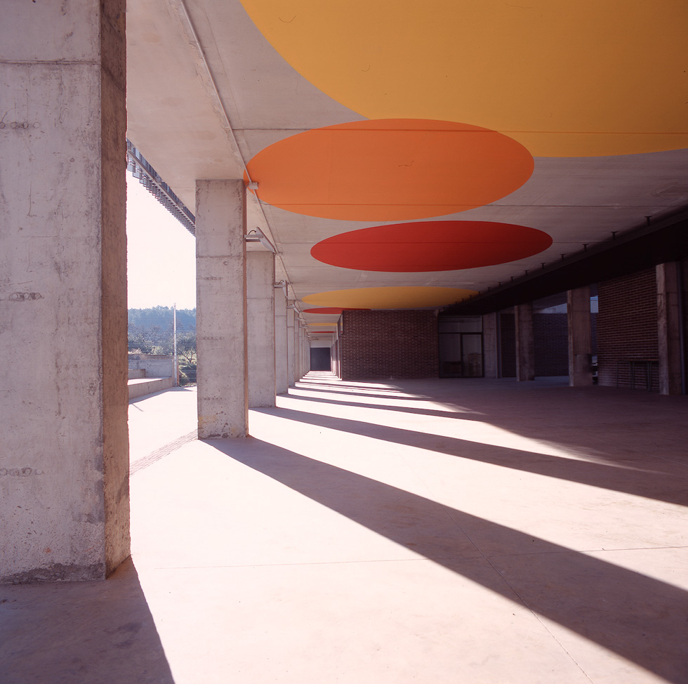 Colorful circular ceiling designs cast shadows on a covered walkway supported by concrete columns.