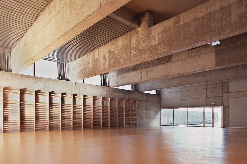 Interior view of a gymnasium with exposed concrete beams and walls, large windows, and wooden gymnastic equipment along one wall.