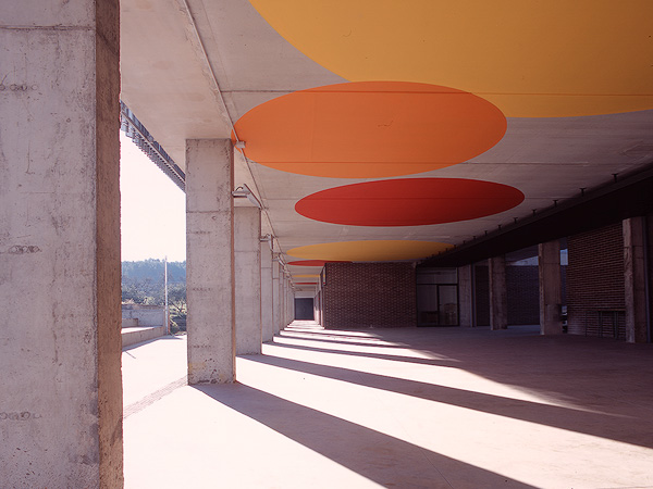 Colorful circular ceiling designs cast shadows on a covered walkway supported by concrete columns.