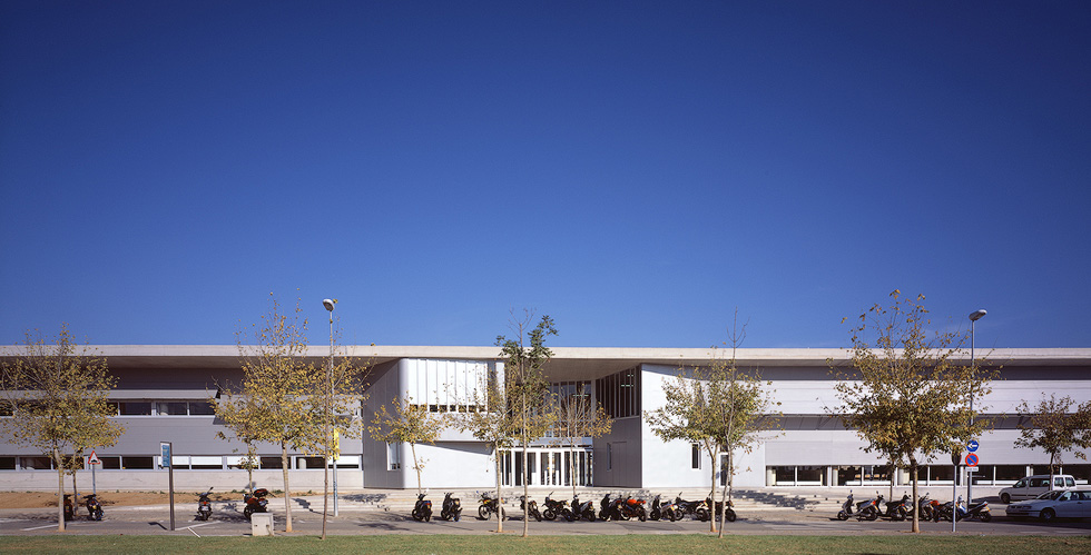 A view of Fontajau High School's modern exterior with a row of trees and parked motorcycles in front, against a clear blue sky.