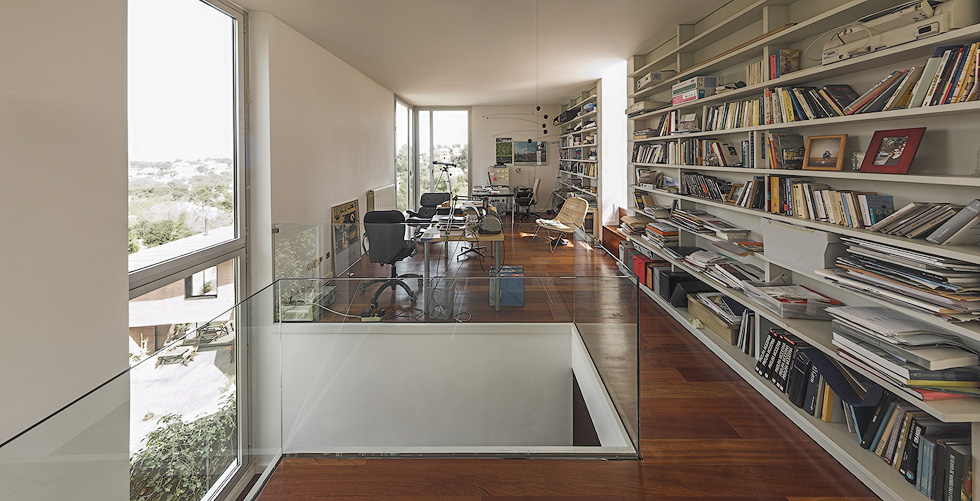 Modern home office with large windows, extensive bookshelves, and wooden floors, overlooking a green landscape.