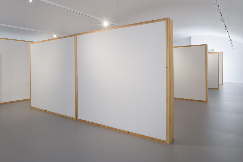 Interior view of a gallery space with white walls and wooden frames, showcasing partition walls for exhibitions.