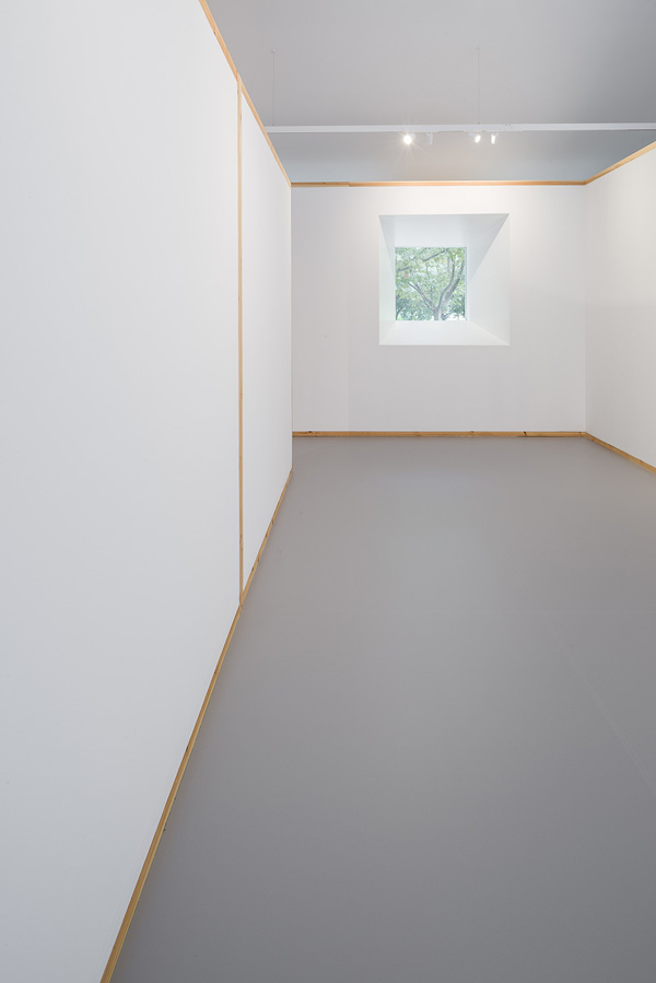 Minimalistic interior with white walls and wooden frames, featuring a single window providing a view of trees outside.