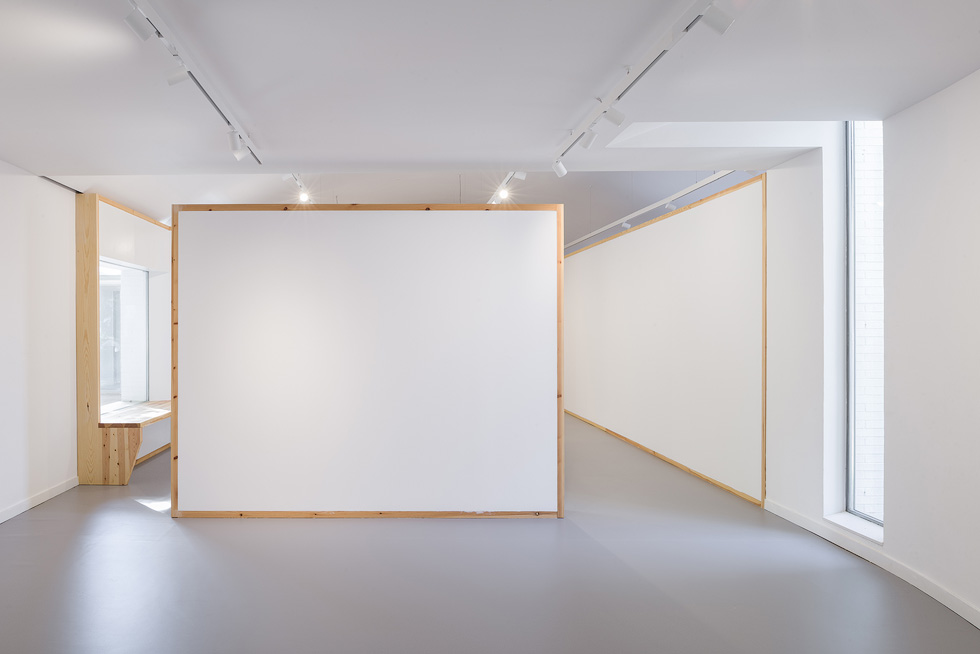 Modern art gallery interior with minimalist design, featuring white walls, wooden frames, and a small seating nook by a large window.